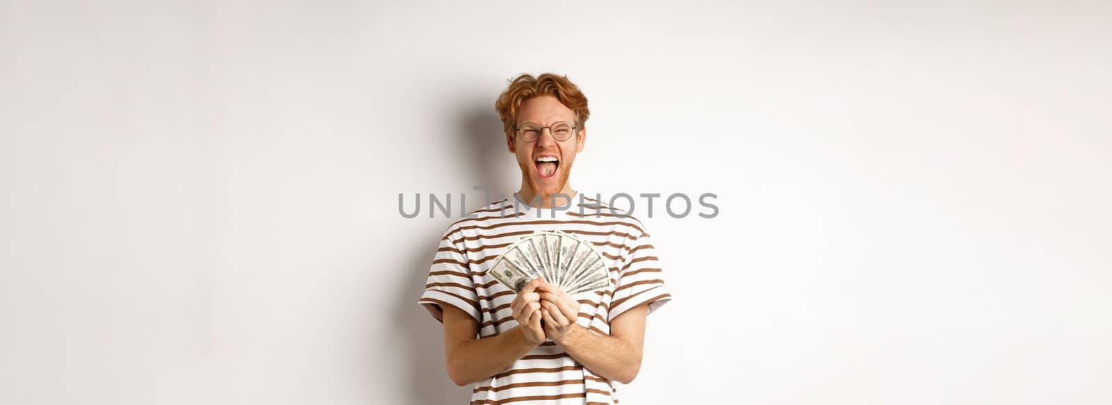 Lucky young man with red hair showing dollars, winning money and screaming of happiness, holding prize cash, standing over white background by Benzoix