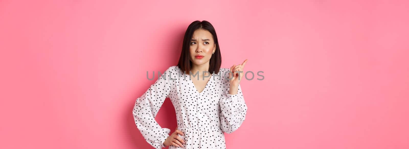Beautiful asian girl pointing finger at upper left corner, looking confused and doubtful, standing over pink background by Benzoix