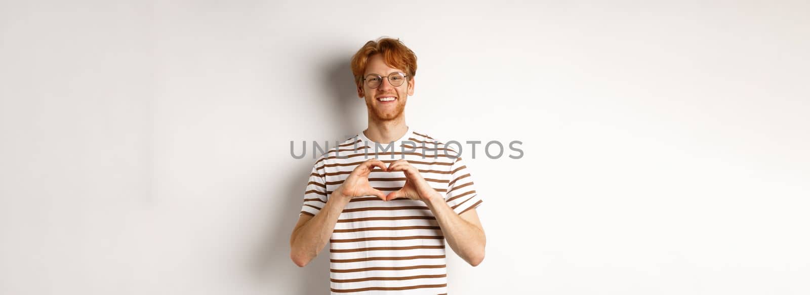 Valentines day. Happy boyfriend with red hair, smiling and showing heart gesture, I love you, standing over white background.