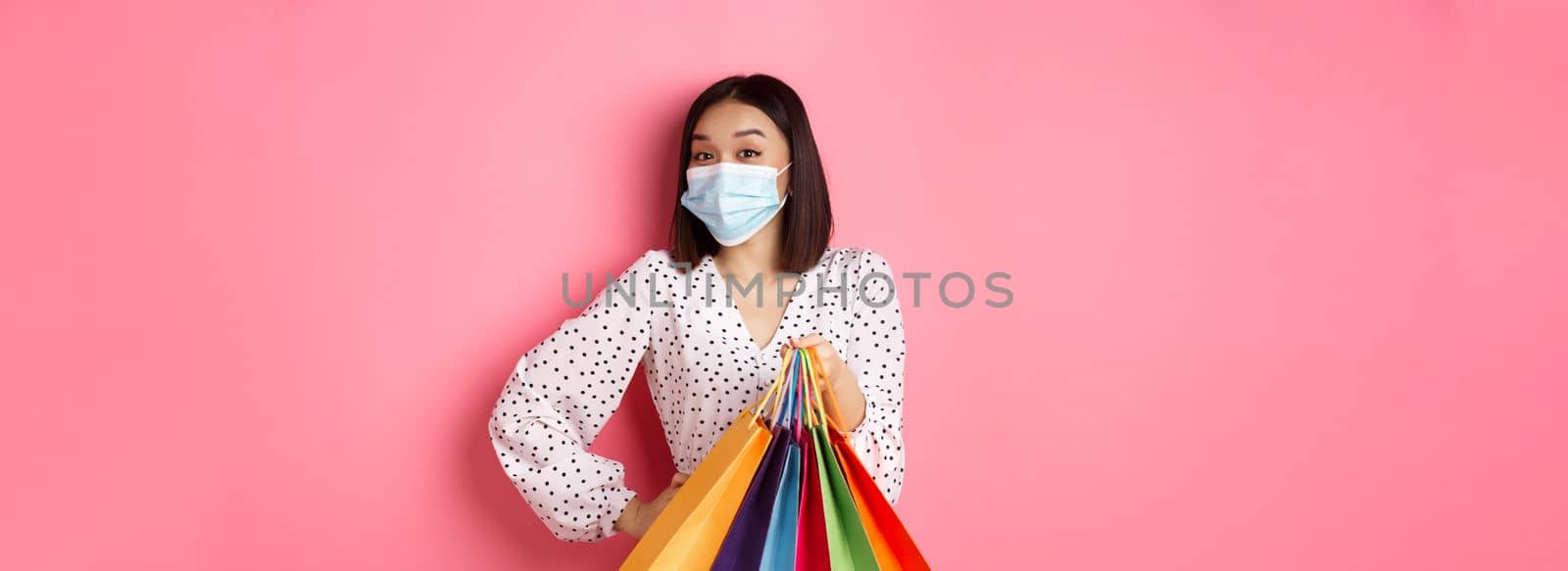 Covid-19, pandemic and lifestyle concept. Beautiful asian woman in medical mask going shopping, holding bags and smiling with eyes, standing over pink background by Benzoix