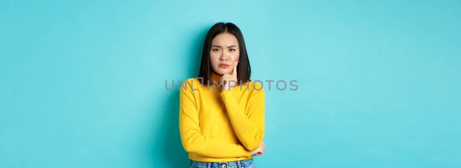 Image of pensive and serious asian woman touching chin, frowning and staring at camera puzzled, making difficult choice, standing over blue background by Benzoix
