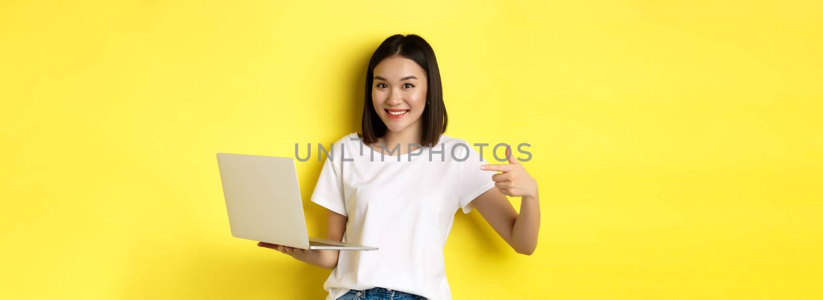 Smiling asian woman pointing finger at her laptop, showing something online, standing over yellow background.