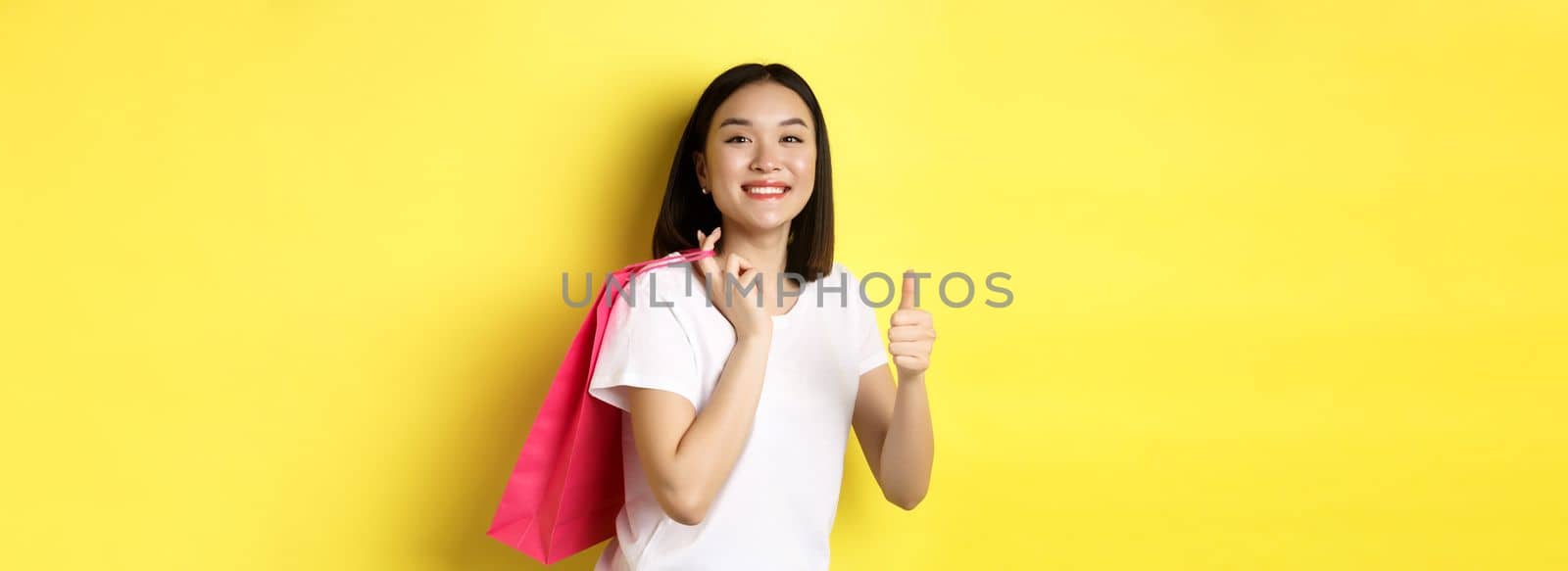 Cheerful asian female shopper looking amused, holding shopping bag and showing thumbs up, recommend store with discounts, standing over yellow background by Benzoix