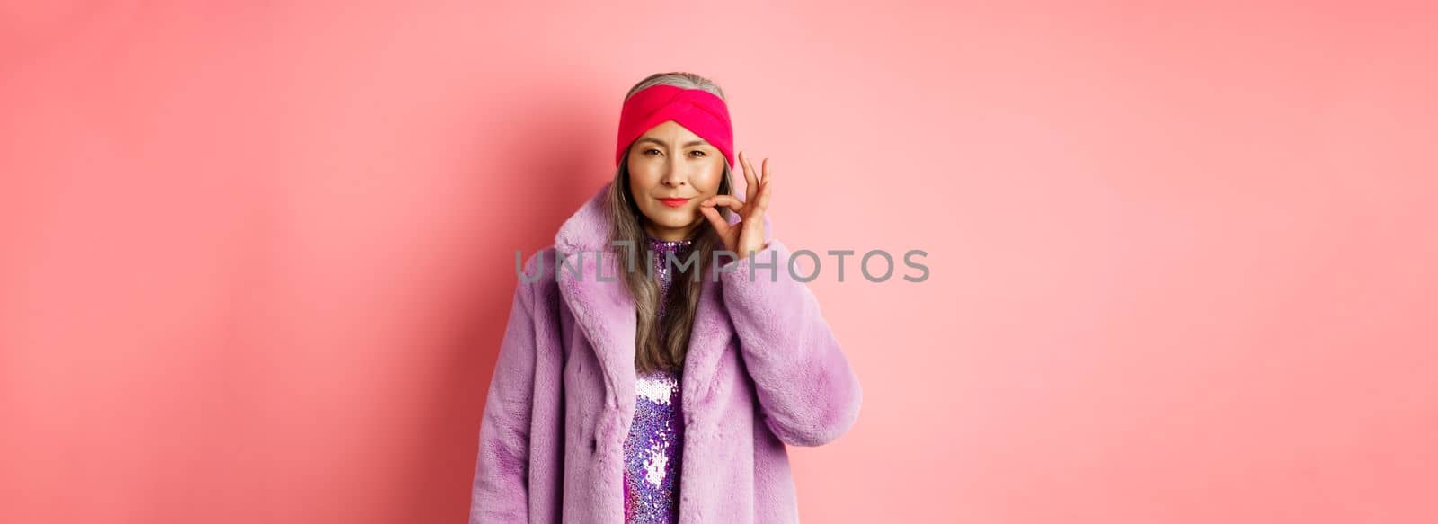 Fashion and shopping concept. Cunning asian senior woman in stylish clothes keeping secret, squinting and looking devious at camera, zipping mouth, making promise, pink background.