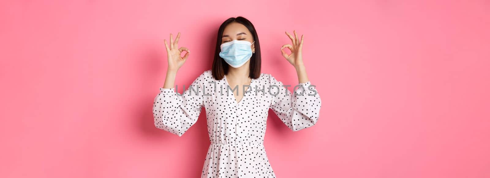 Coronavirus, social distancing and lifestyle concept. Satisfied asian woman in face mask showing okay signs, approve and agree, praising good choice, pink background.
