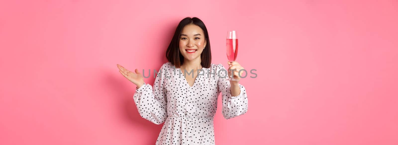 Beautiful asian woman raising glass of champagne and looking happy at camera, congratulating you on party, celebrating, standing over pink background.