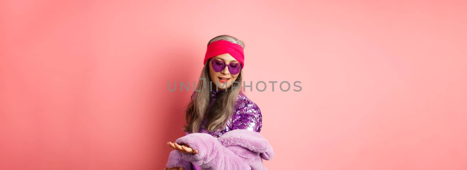 Fashionable mature asian woman in glittering dress dancing on party, having fun and relaxing, standing over pink background.