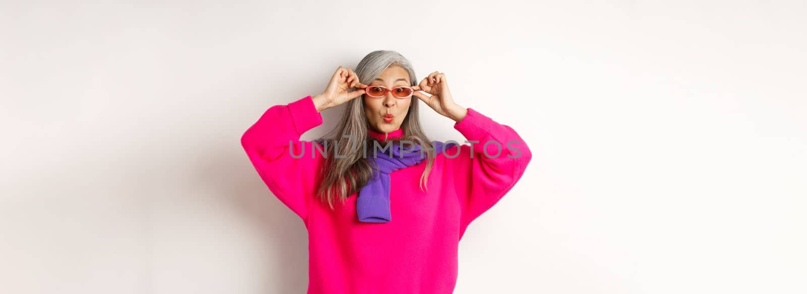 Trendy asian senior woman in sunglasses, pucker lips and looking surprised at camera, staring amazed at special offer, standing over white background.