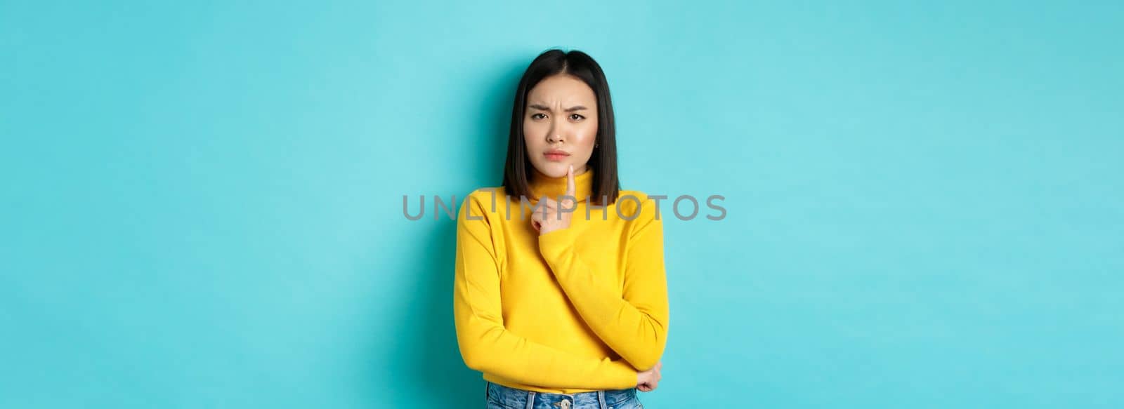 Image of pensive asian woman touching lip and frowning, thinking about something, trying to understand, standing over blue background by Benzoix