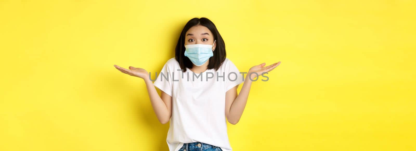Covid-19, quarantine and social distancing concept. Clueless asian woman in medical mask shrugging shoulders, spread hands sideways clueless, know nothing, yellow background.