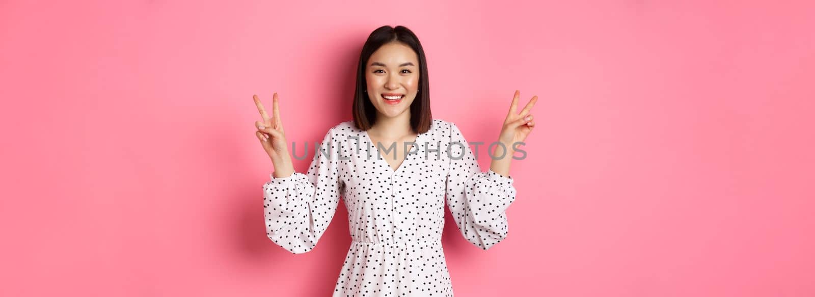 Cute asian brunette girl in dress smiling, showing kawaii peace signs and looking happy, standing over pink background.