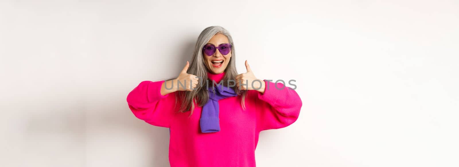 Shopping concept. Stylish asian senior woman in sunglasses and trendy outfit, showing thumb-up in approval, recommending shop, standing over white background by Benzoix