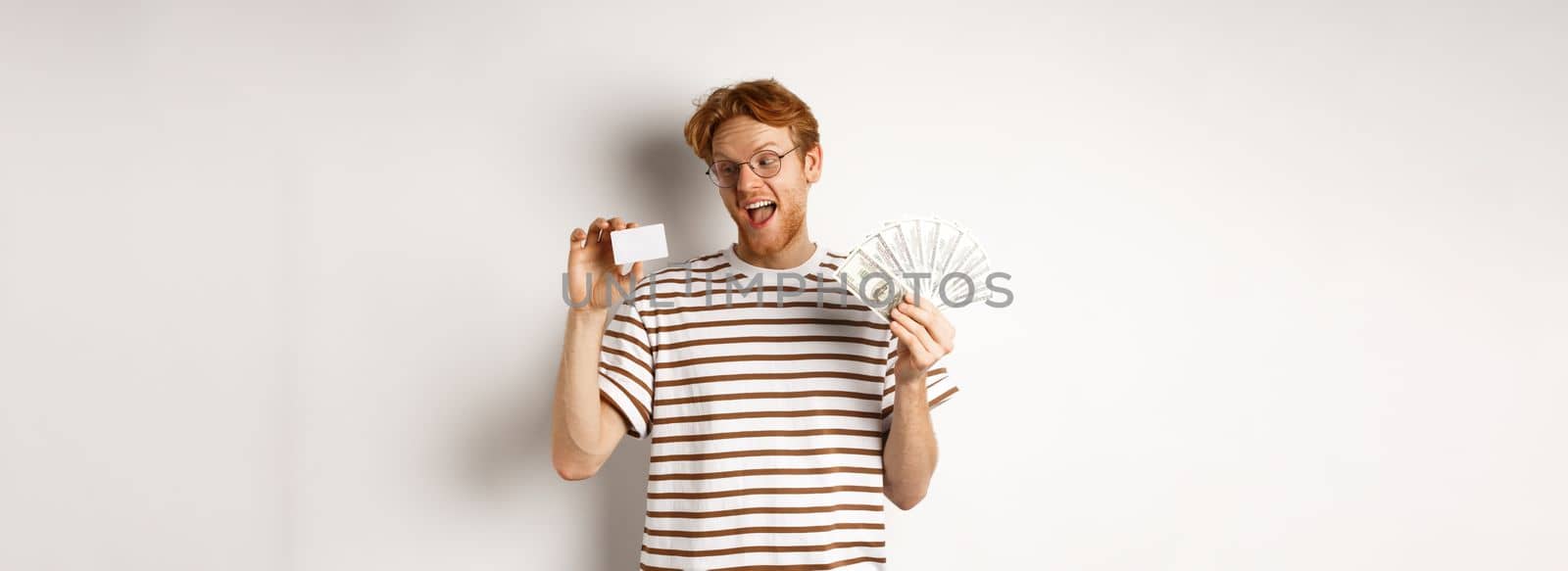 Shopping and finance concept. Young redhead man in glasses choosing plastic credit card, showing dollars and smiling, standing over white background.