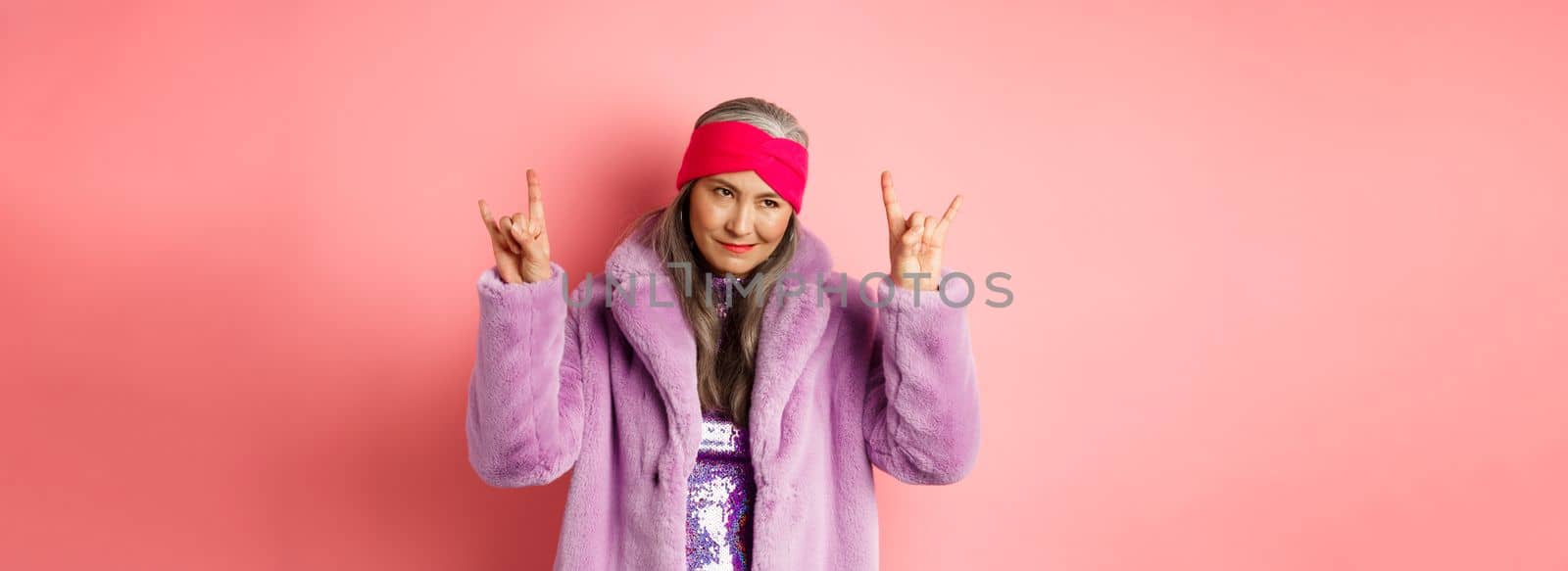 Funny and cool asian senior woman showing rock n roll gesture, looking sassy, standing over pink background.