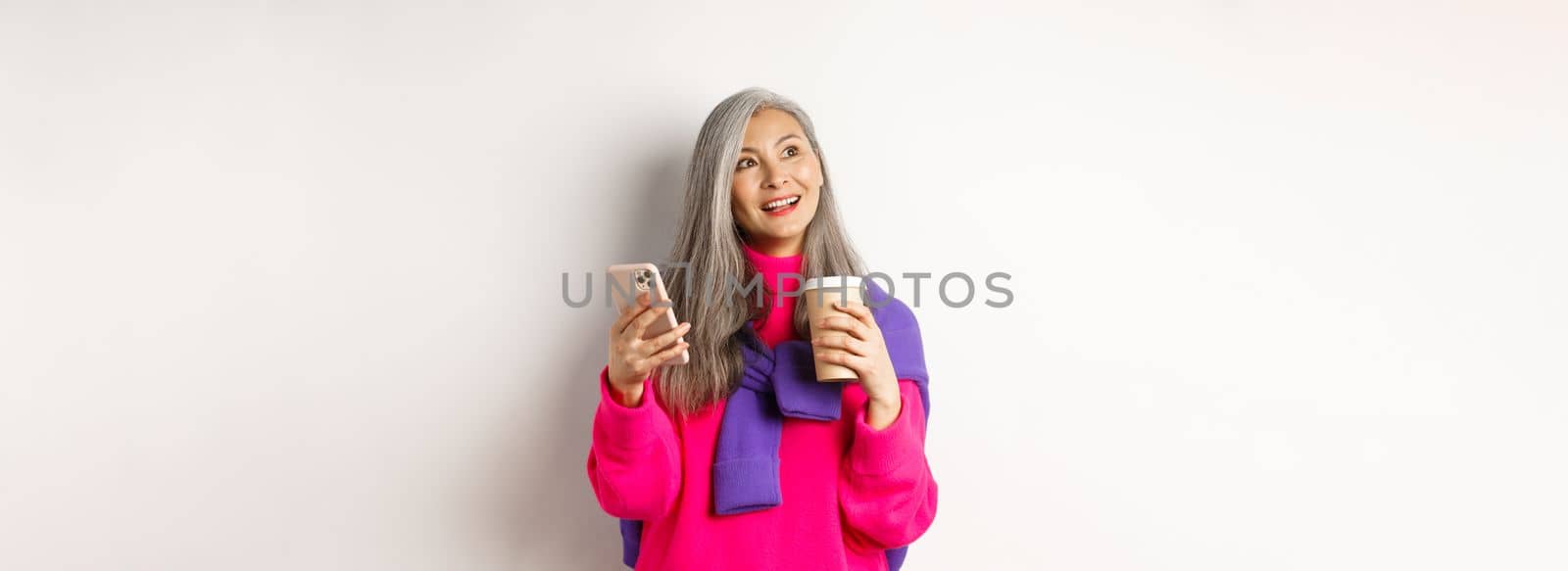 Fashionable asian mature woman with grey hair, drinking coffee and using smartphone, looking upper left corner promotion with dreamy face, white background by Benzoix