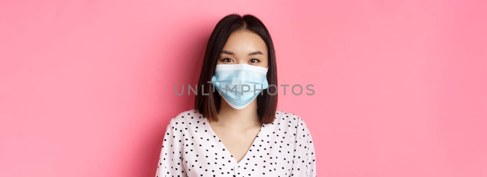 Covid-19, pandemic and lifestyle concept. Close-up of beautiful asian woman in face mask smiling with eyes, wearing protection from coronavirus in public, pink background.