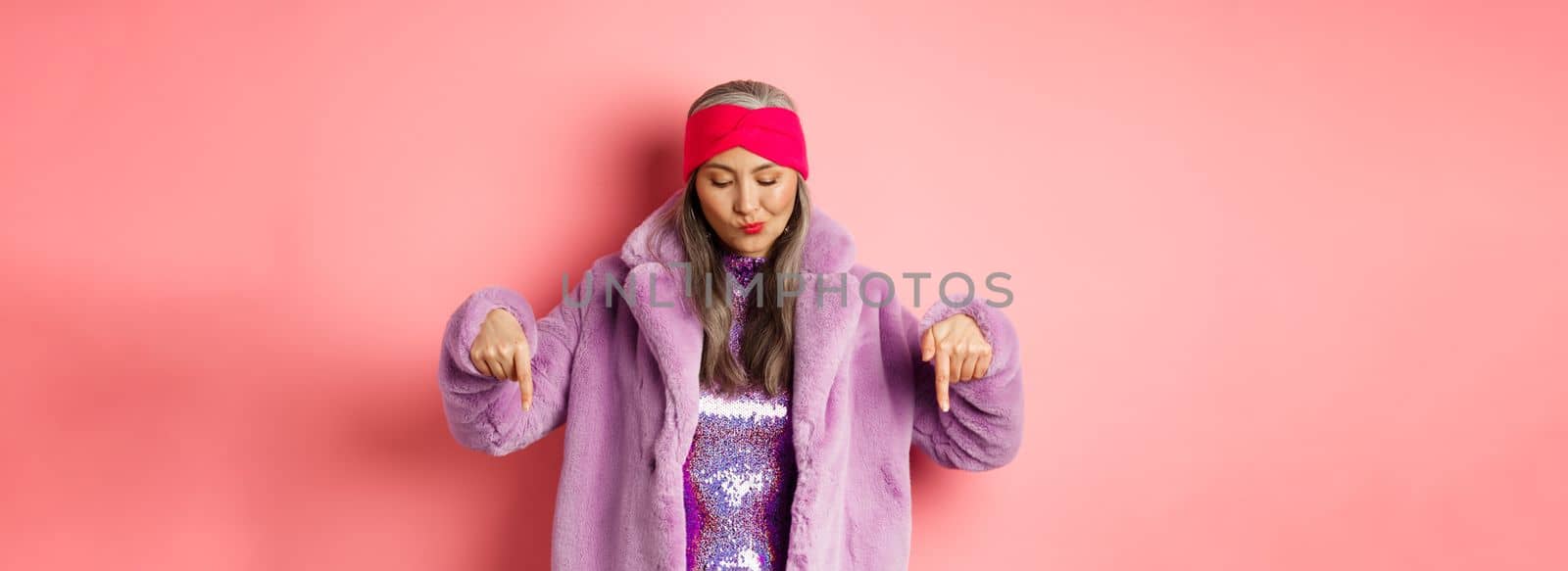 Fashion and shopping concept. Funky asian senior woman in trendy faux fur coat and disco dress, looking and pointing down, standing over pink background.