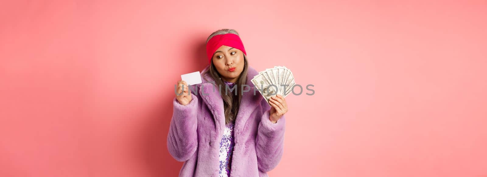 Shopping and fashion concept. Thoughtful asian female holding dollars but thinking about paying contactless with plastic credit card.