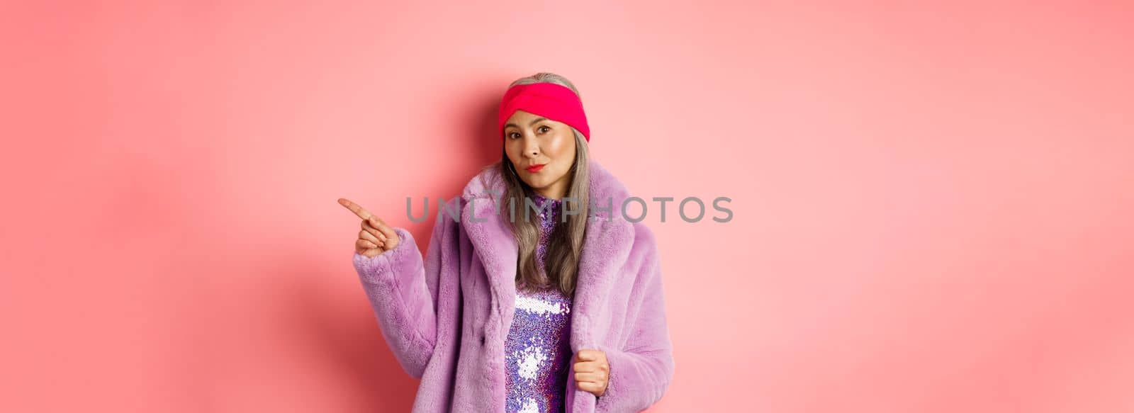 Trendy asian senior woman in cool purple winter coat pointing finger right, showing advertisement and looking sassy at camera, pink background.