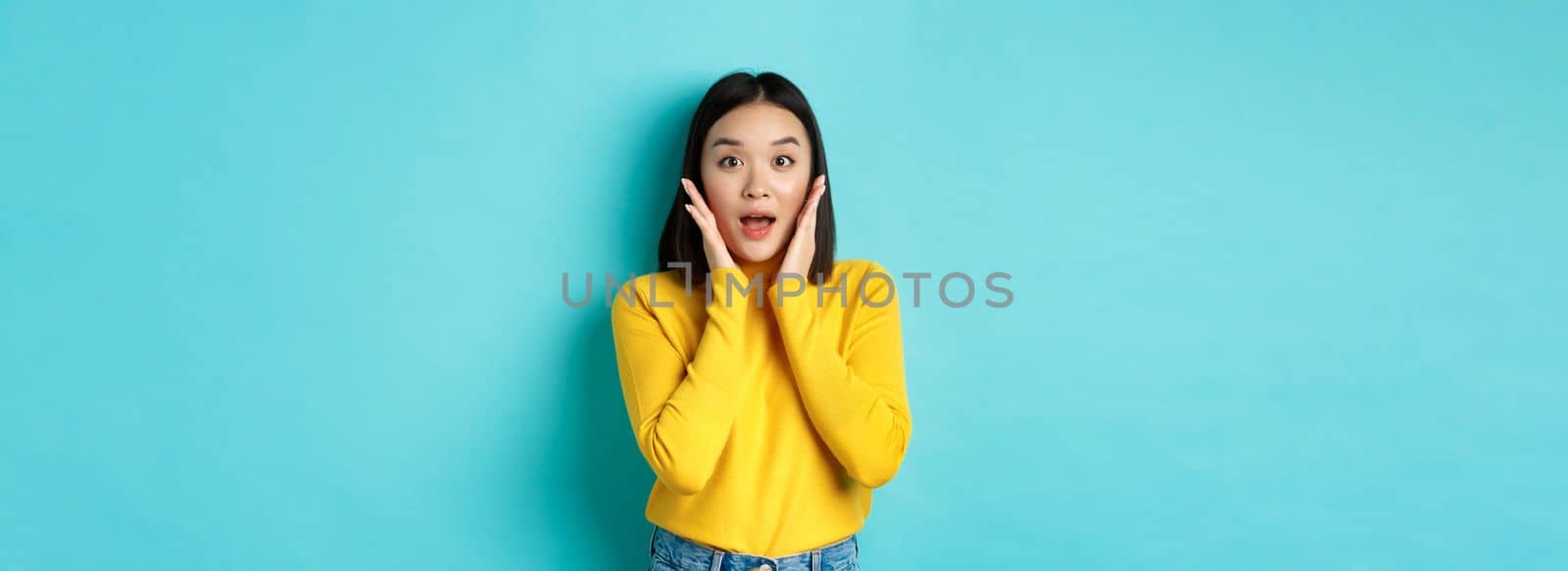 Portrait of surprised asian girl checking out promo, gasping amazed and touching cheeks, staring at camera amazed, blue background by Benzoix