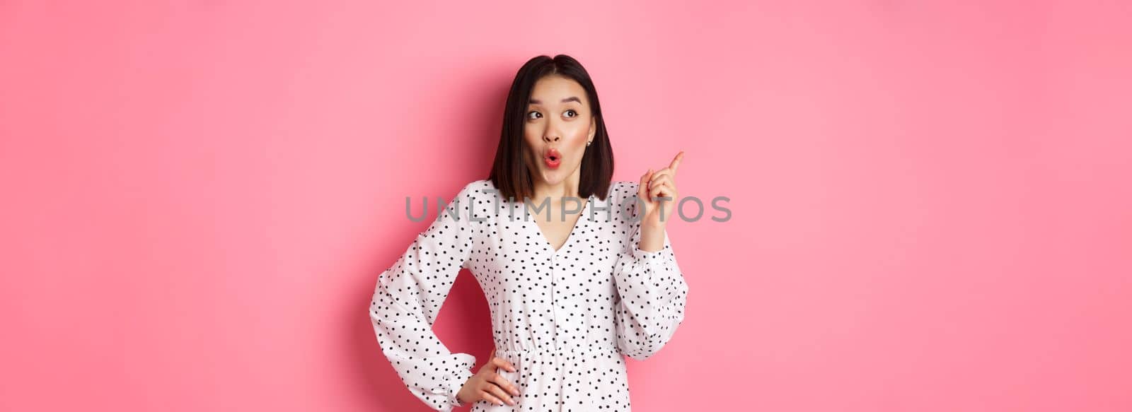 Amazed asian girl in dress pointing finger at upper left corner promo offer, looking interested, shopping and standing over pink background by Benzoix