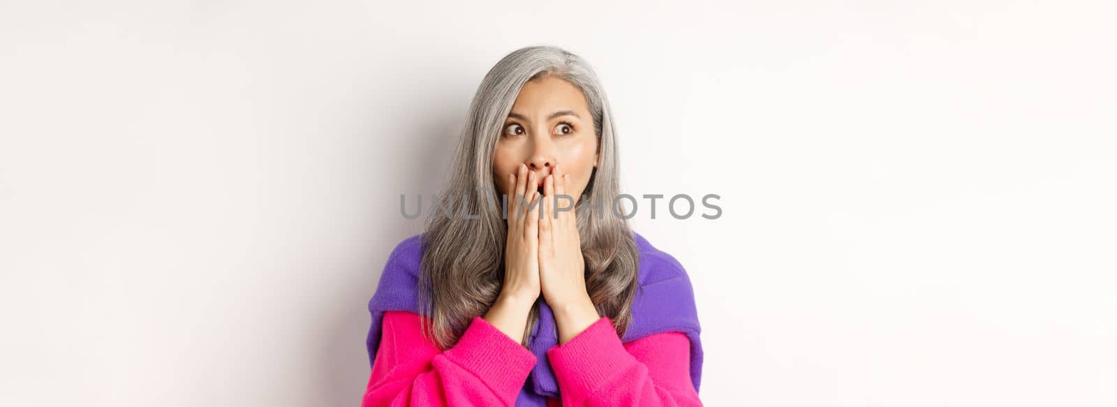 Close-up of shocked asian middle-aged woman with grey hair, gasping and covering mouth startled, looking left, realising something, white background.