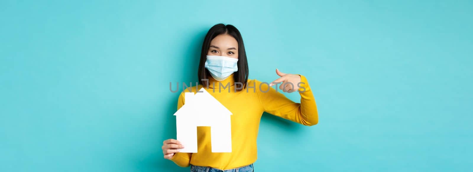 Covid-19, pandemic and real estate concept. Cheerful asian woman smiling in medical mask, showing paper house cutout, recommend agency for buying property, blue background by Benzoix