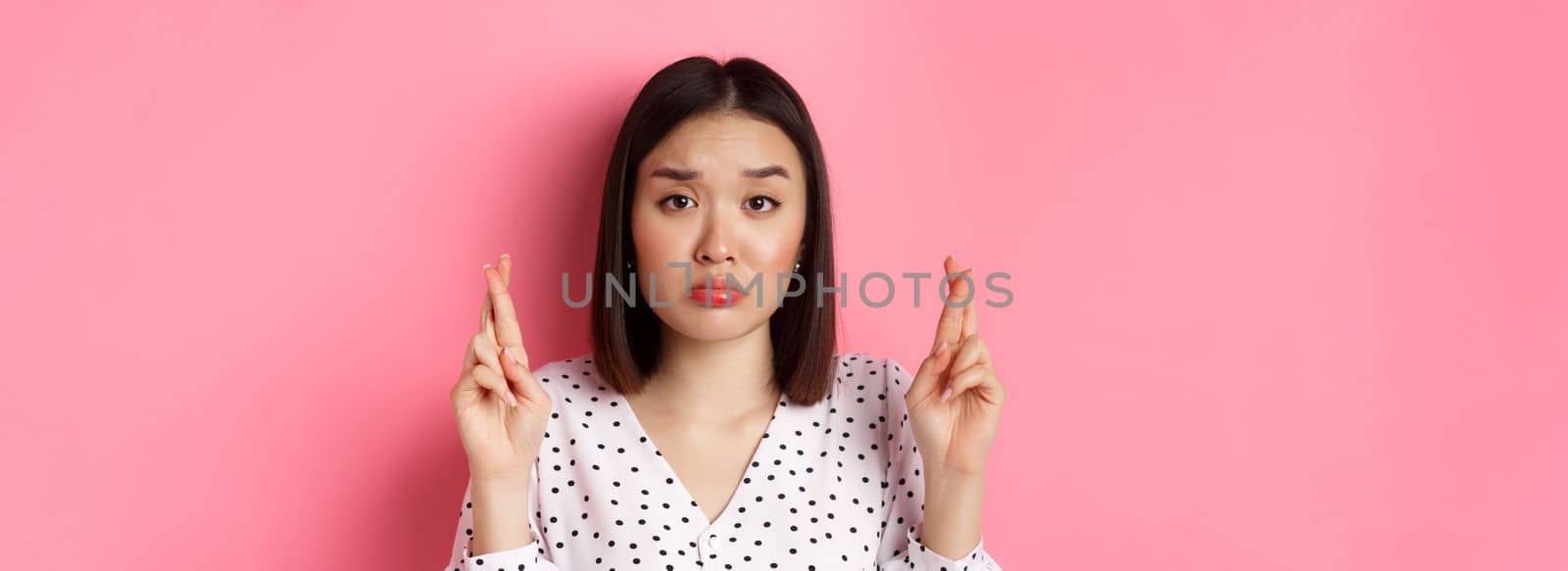 Beauty and lifestyle concept. Close-up of sad and hopeful asian woman making wish, cross fingers good luck and pouting, looking upset, standing over pink background.