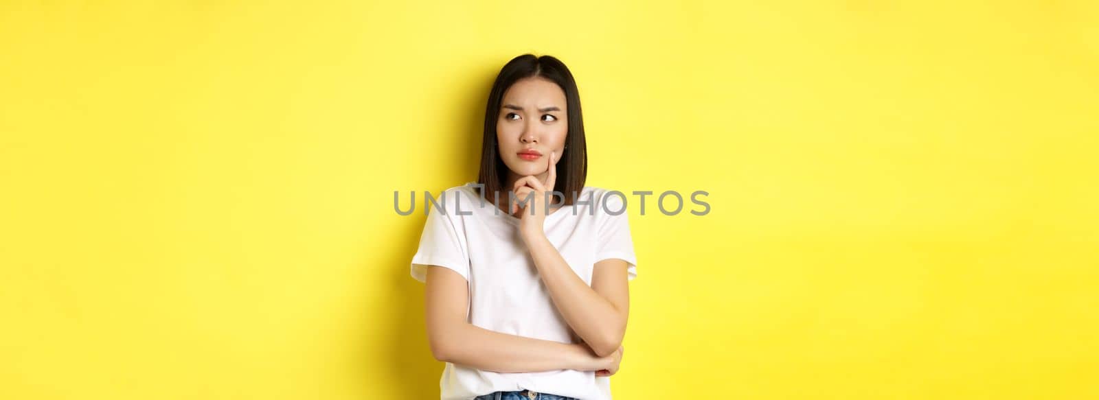 Beauty and fashion concept. Pensive asian woman thinking, looking thoughtful while pondering something, standing over yellow background.