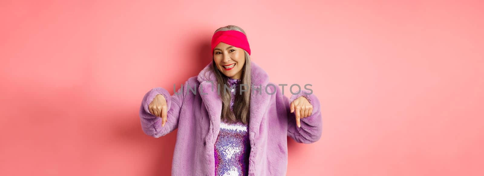 Fashion and shopping concept. Smiling senior asian woman pointing fingers down, showing store advertisement on bottom, wearing stylish purple winter coat, pink background by Benzoix