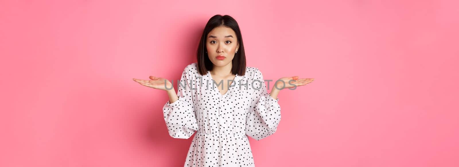 Indecisive cute asian woman shrugging, holding hands spread sideways on copy spaces, dont know, having doubts, standing over pink background.