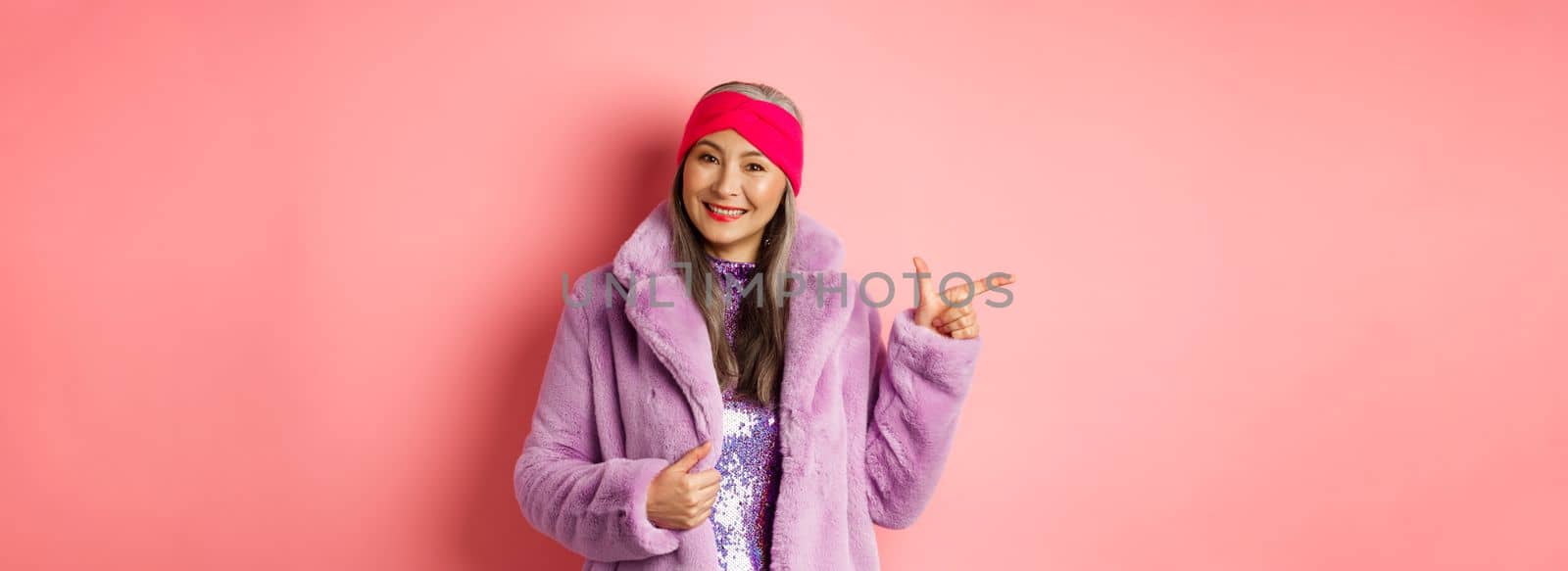 Fashion and shopping concept. Stylish senior asian woman pointing finger left at promotion deal, smiling at camera, suggesting special offer, standing against pink background.
