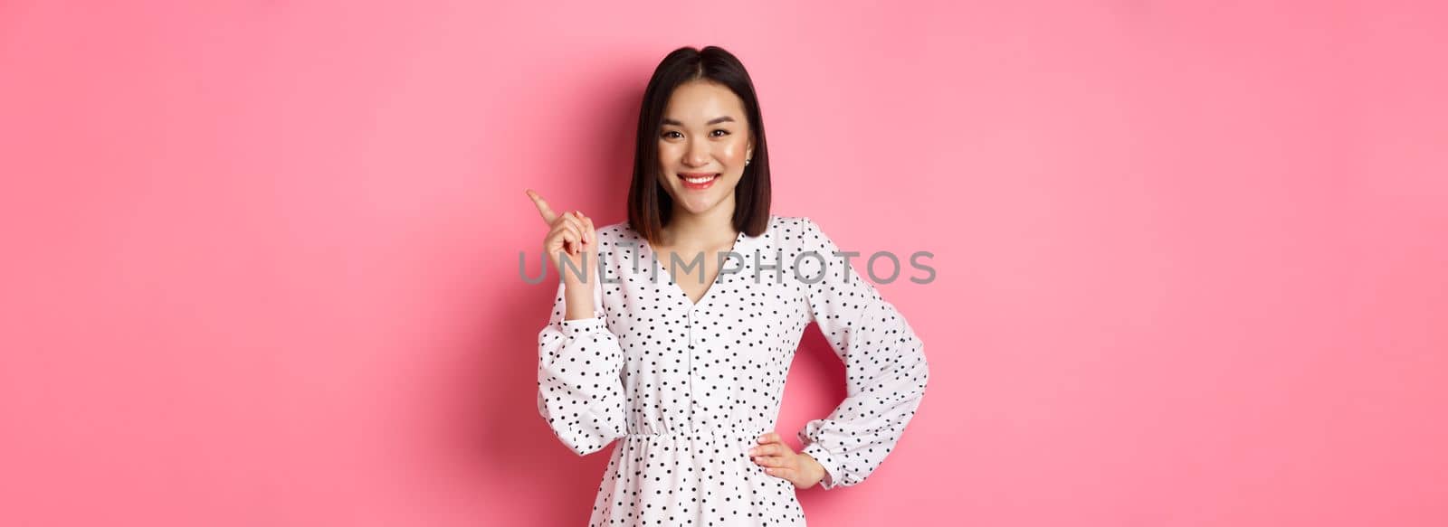 Beautiful asian female model smiling, pointing finger at upper right corner copy space, showing advertisement banner, standing over pink background.