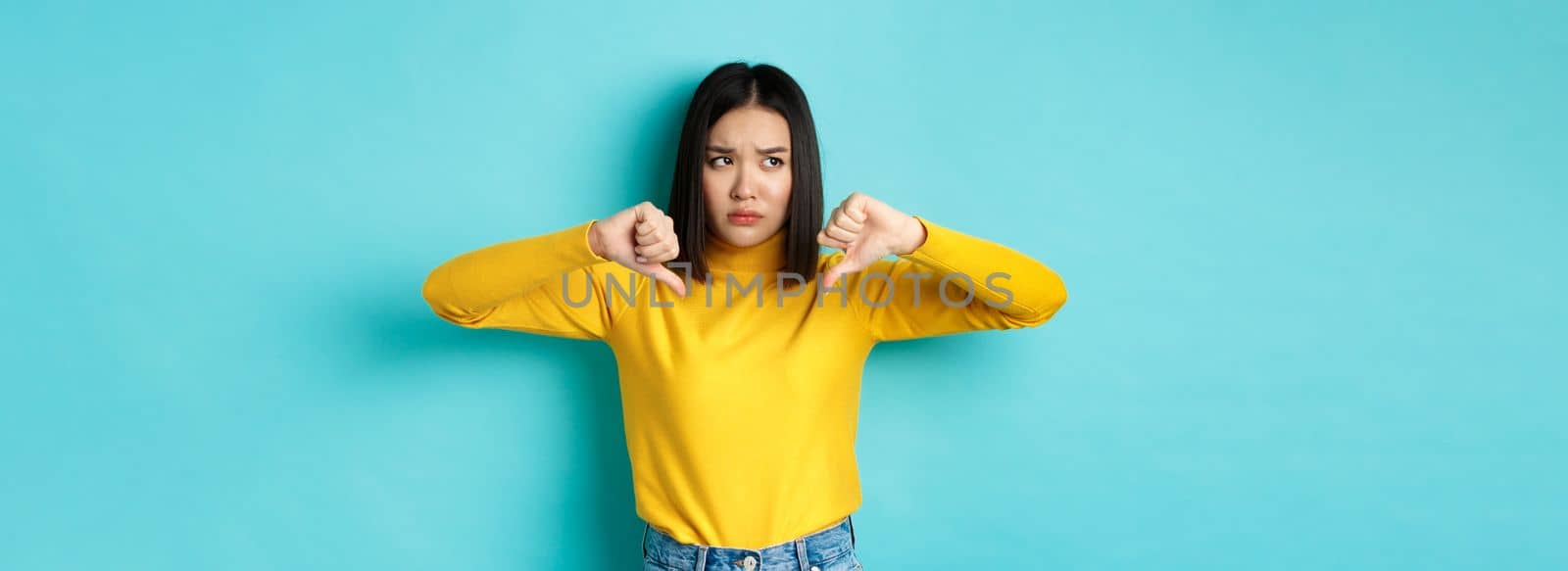 Disappointed asian woman frowning upset, showing thumbs down in dislike and disapproval, standing over blue background, looking left.
