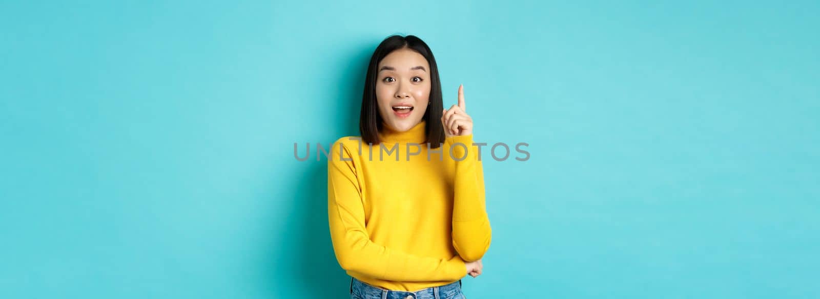 Excited asian woman with short dark hair, pitching an idea, raising finger in eureka gesture and smiling, standing over blue background.