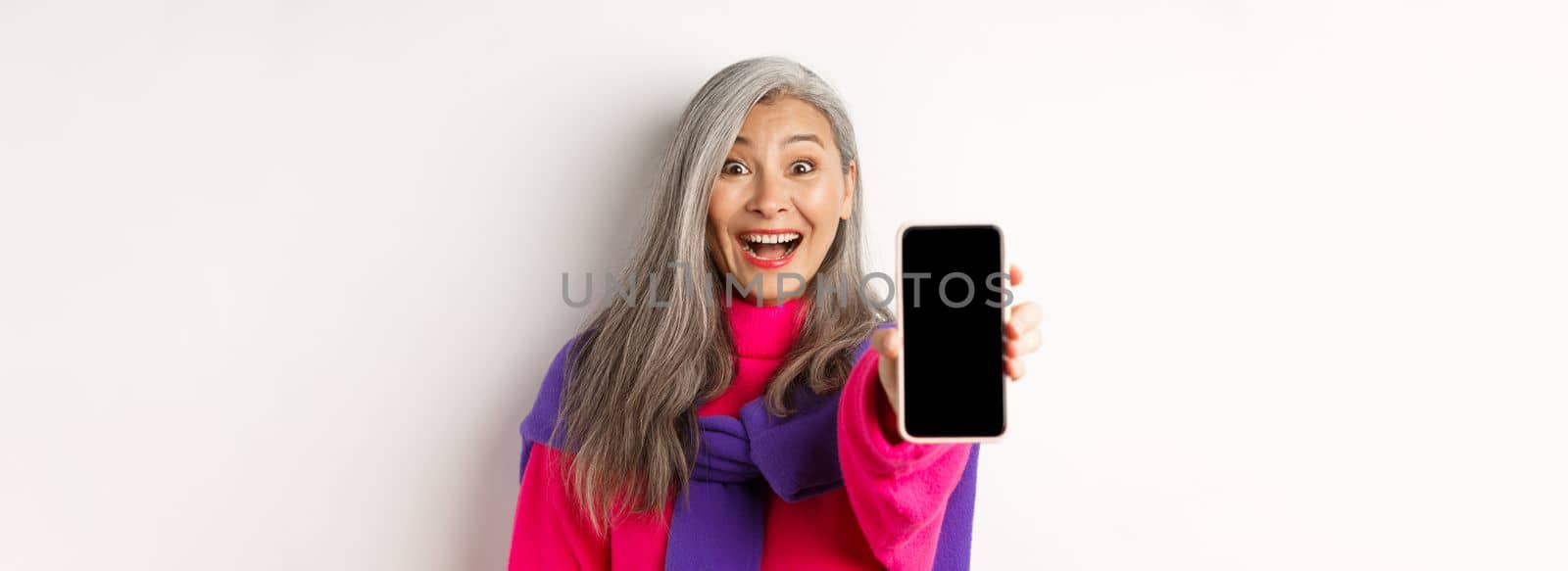 Online shopping. Close up of stylish asian senior woman extending hand with mobile phone, showing blank smartphone screen and smiling, standing over white background.