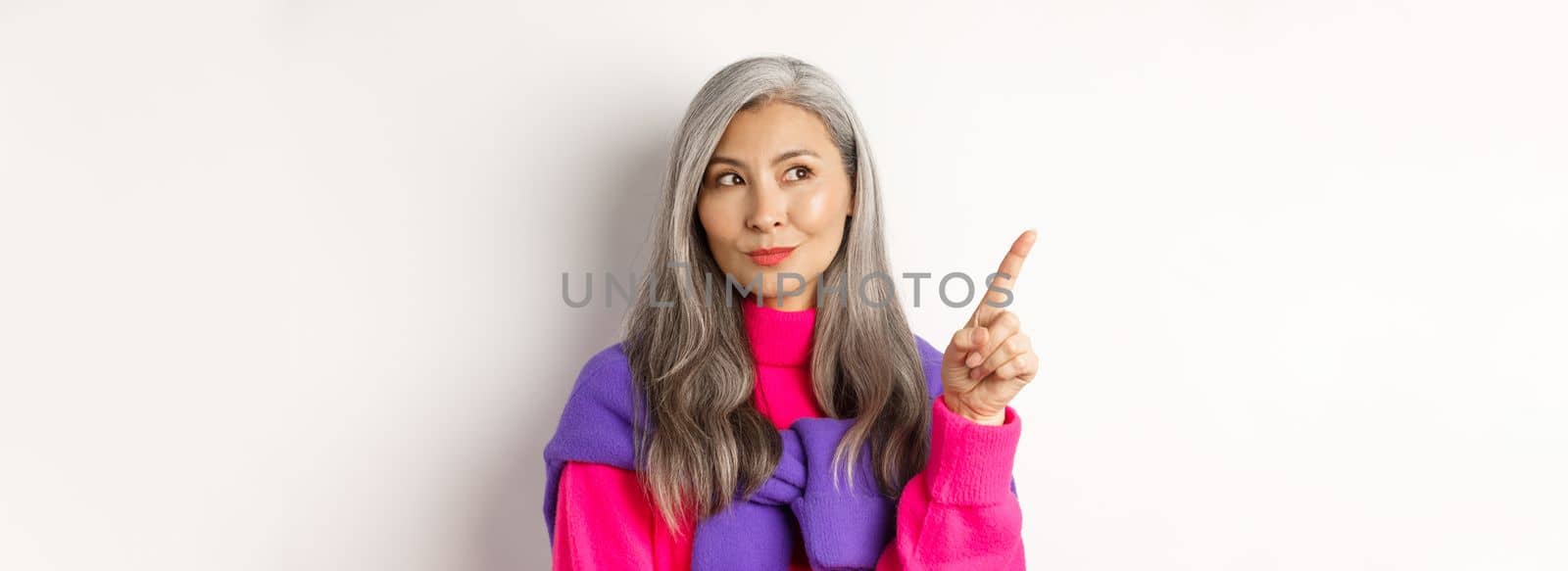 Close-up of stylish korean senior woman with grey hair looking and pointing upper left corner, showing promotion offer, standing over white background by Benzoix