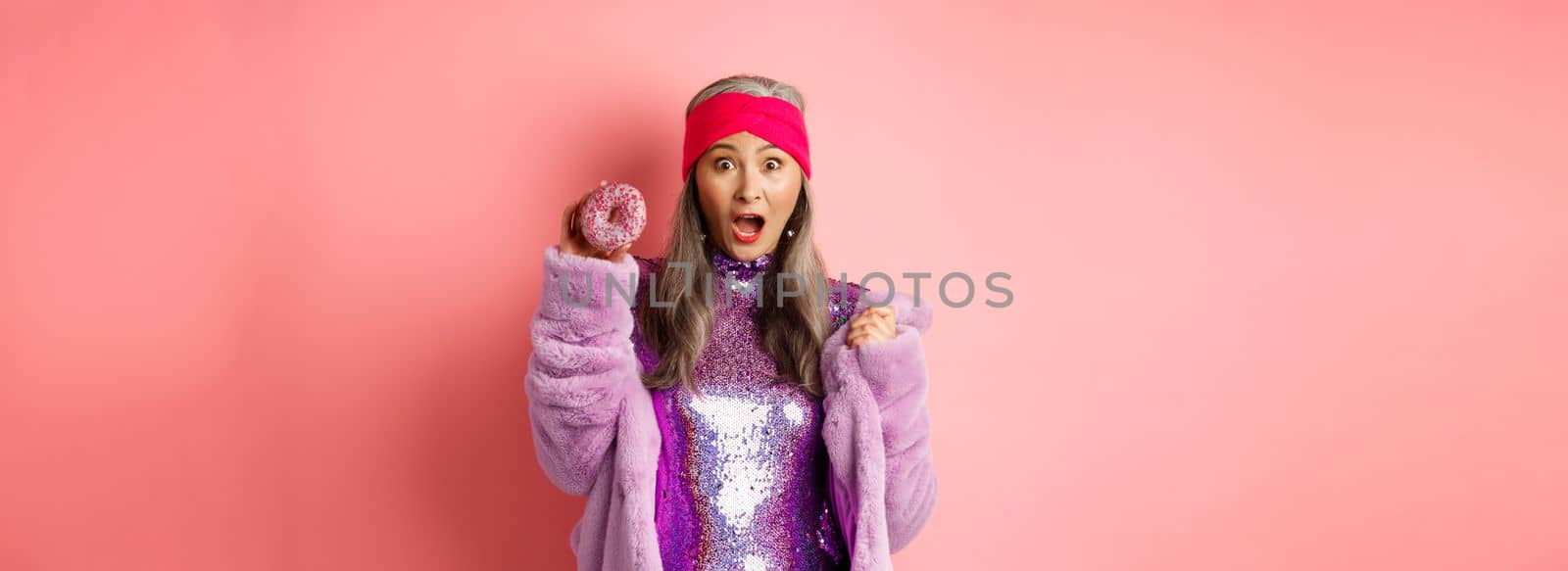 Fashion and food concept. Beautiful asian senior woman in stylish glittering dress holding sweet donut and screaming at camera, scared of calories, pink background by Benzoix