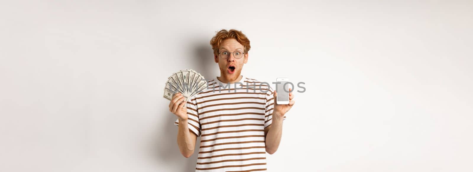 Amazed redhead man showing smartphone app on blank screen and money, winning prize cash online, standing over white background by Benzoix