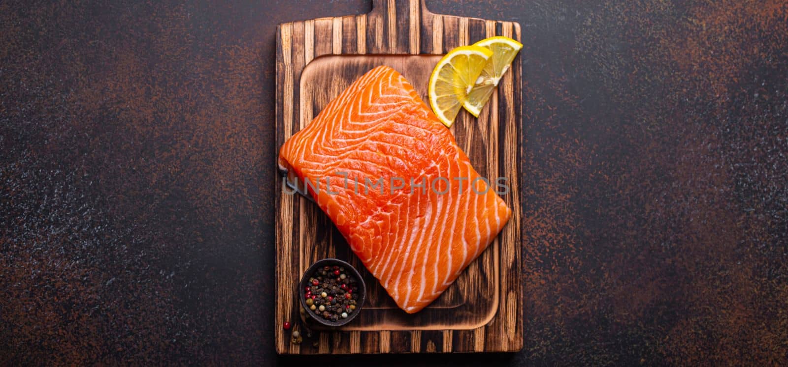 Fresh raw salmon fillet on wooden kitchen cutting board with seasonings and lemon top view on dark brown rustic background