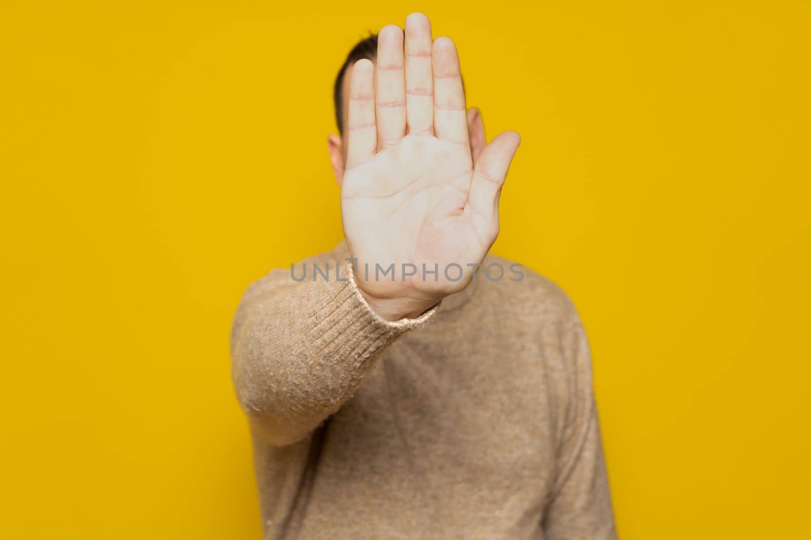 Hispanic man putting hand in front to stop camera, rejecting photos or images against yellow colored wall