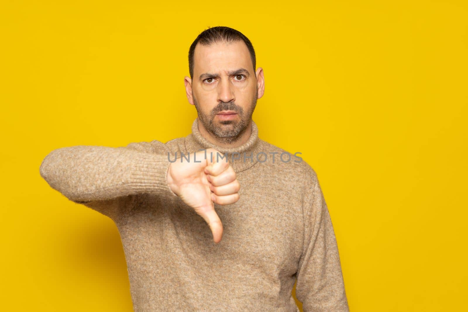 Hispanic man with a beard wearing a beige turtleneck posing with a serious expression over a yellow background has his thumbs down in disapproval