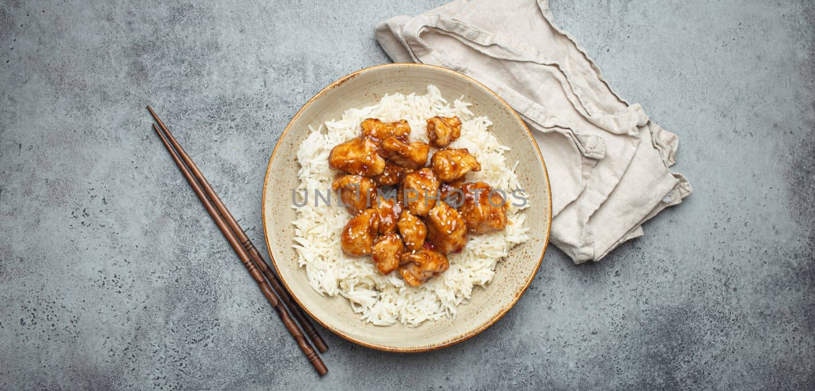 Chinese sweet and sour sticky chicken with sesame seeds and rice on ceramic plate with chopsticks top view, gray rustic stone background, traditional dish of China by its_al_dente