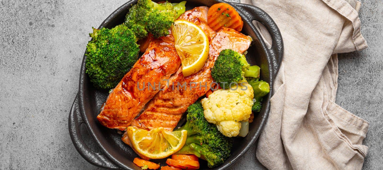 Healthy baked fish salmon steaks, broccoli, cauliflower, carrot in black cast iron casserole bowl on grey rustic stone background. Cooking a delicious low carb dinner, healthy nutrition concept by its_al_dente