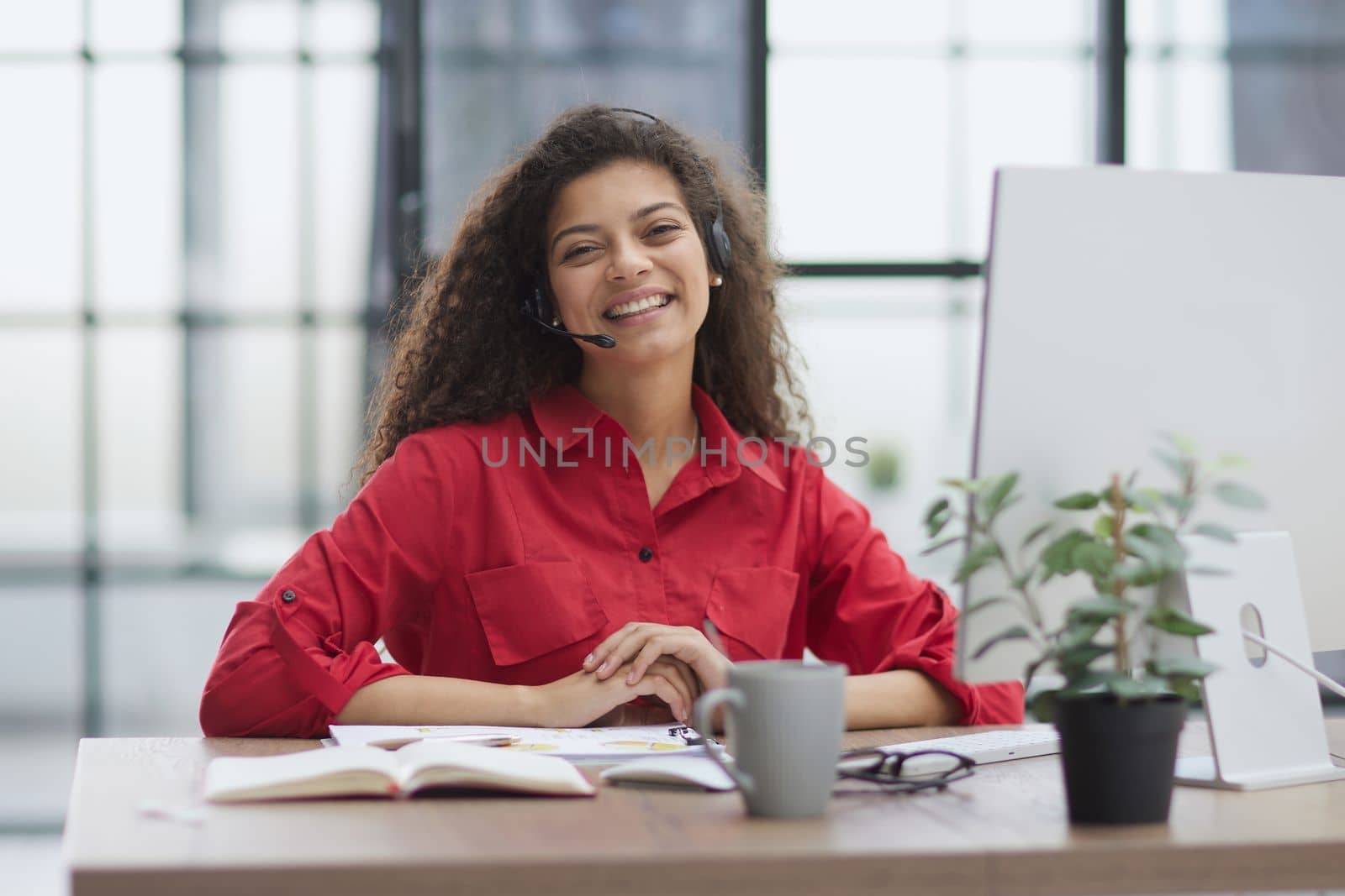 woman in a red shirt sits at a table in a red jacket in headphones with a microphone.