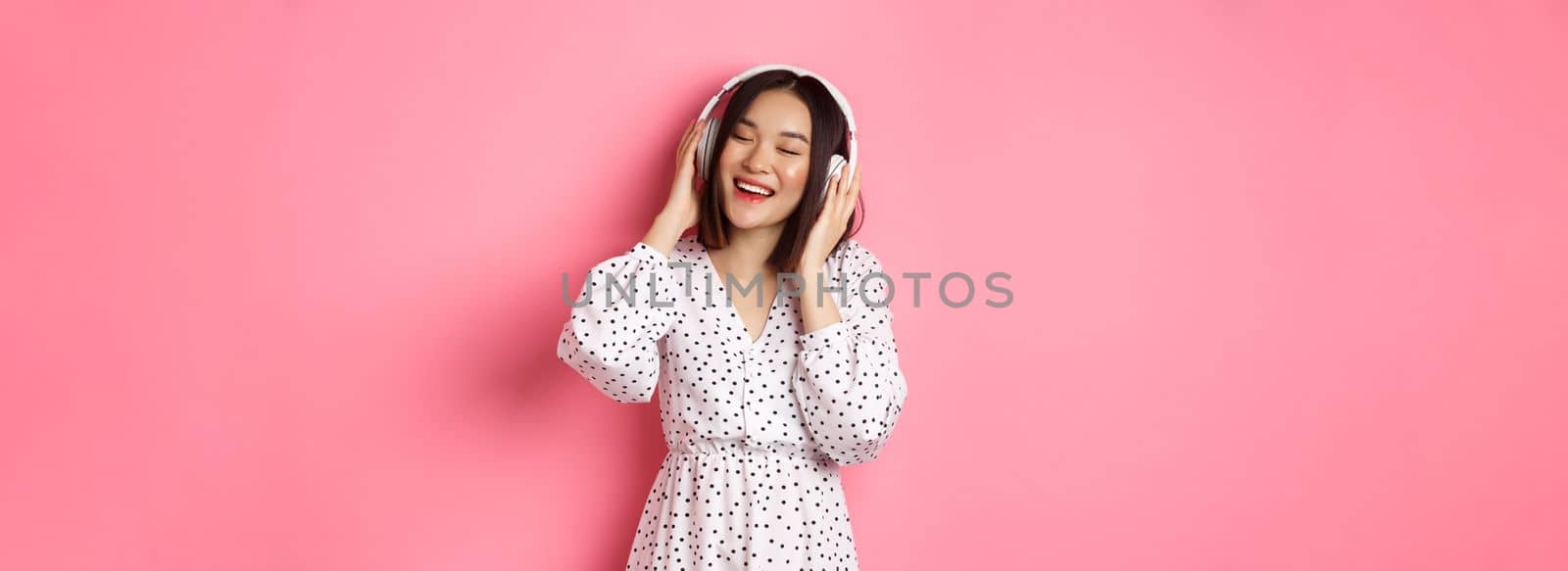 Romantic asian woman smiling happy, listening music in headphones and dancing, standing in trendy dress over pink background.