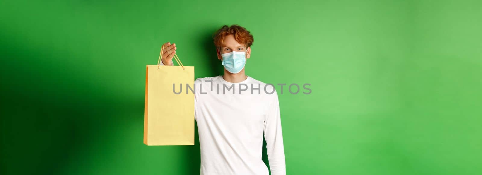 Portrait of handsome boyfriend in face mask giving shopping bag with valentines gift, smiling with eyes, standing over green background.