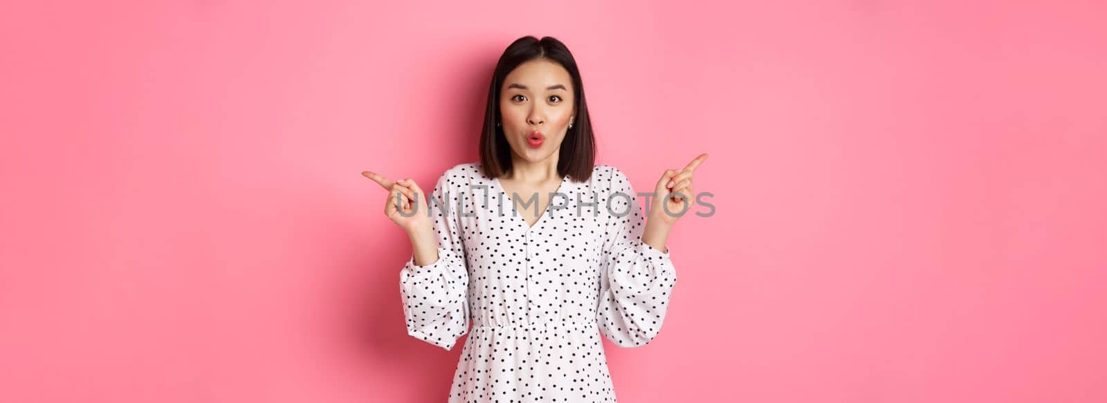 Beautiful asian woman making choice on shopping, pointing fingers sideways and showing variants, smiling at camera, standing over pink background.