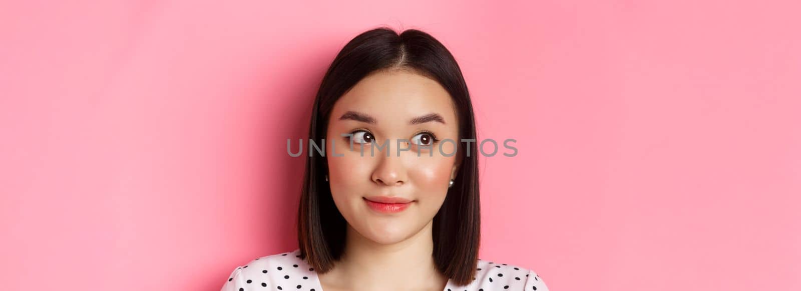 Beauty and skin care concept. Close-up of cute asian teenage girl looking left at banner, smiling silly, standing in dress over pink background.