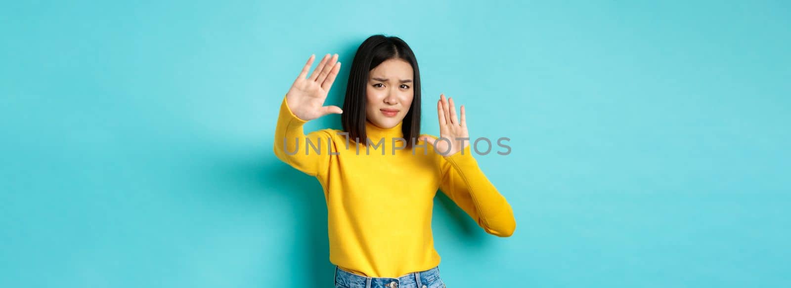 Image of woman feeling uncomfortable in front of camera, cover face with hands and frowning, begging to stop, standing against blue background by Benzoix